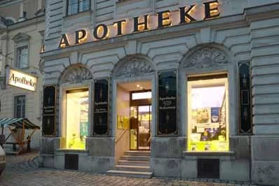 Eingang der Schottenapotheke am Abend mit goldenem Schriftzug „APOTHEKE“ und beleuchteten Schaufenstern